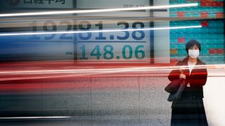 In this April 24, 2020, file photo, a woman wearing a mask to help stop the spread of the new coronavirus stands near an electronic stock board showing Japan's Nikkei 225 index at a securities firm in Tokyo.