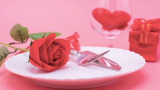 Valentines day dinner with table place setting with red gift, glass for champagne, a bottle of champagne, heart ornaments with silverware on white background. Close up. Valentine’s card