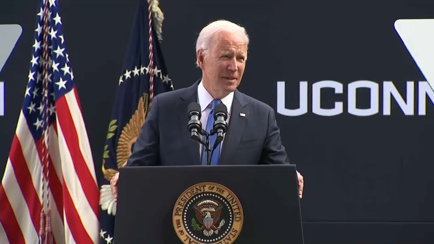 President biden stands at a podium with flags on his right side and a UConn logo on the left.