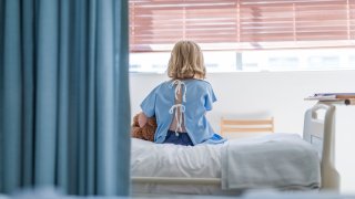Rear view of sick girl with stuffed toy sitting on bed. Female child patient is resting in ward at clinic. She is wearing hospital gown.