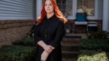 A photo of Abigail Zwerner. She has red hair and is wearing a black blouse, standing against the sun in front of a white house.