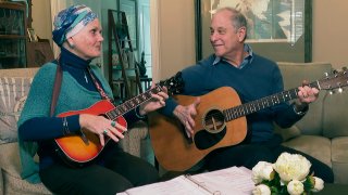 A photo of Lynda Shannon Bluestein on the left, playing the guitar with her husband Paul in the living room of their home, Feb. 28, 2023, in Bridgeport, Connecticut. Both are wearing blue sweaters, with Lynda wearing a blue headband.