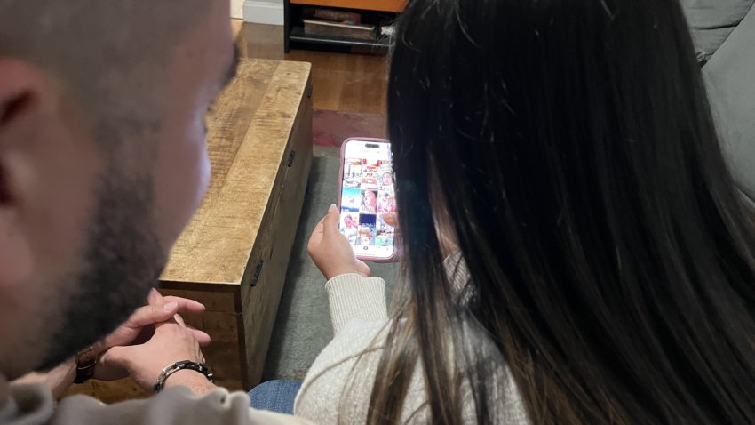 A man and a woman with long hair sit on a couch and look at a phone where the TikTok social media app is visible