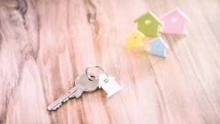 Silver Key with Breloque in Form of House and some Miniature Symbol of Houses on Side in Different Colours on Wooden Surface