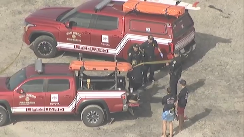 A small group of lifeguard vehicles was on the scene when Sky Ranger 7 flew overhead around 2:20 p.m., surrounded by caution tape, with a yellow tarp covering what was presumably a body on the beach between the trucks.