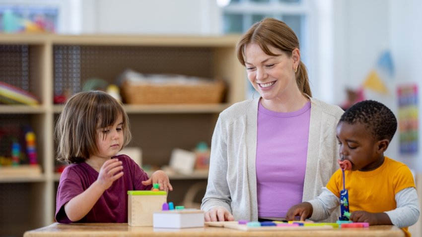 Two daycare children stand at a table together as they play with various toys. Their teacher sits with them as she helps lead and instruct their play to facilitate learning.