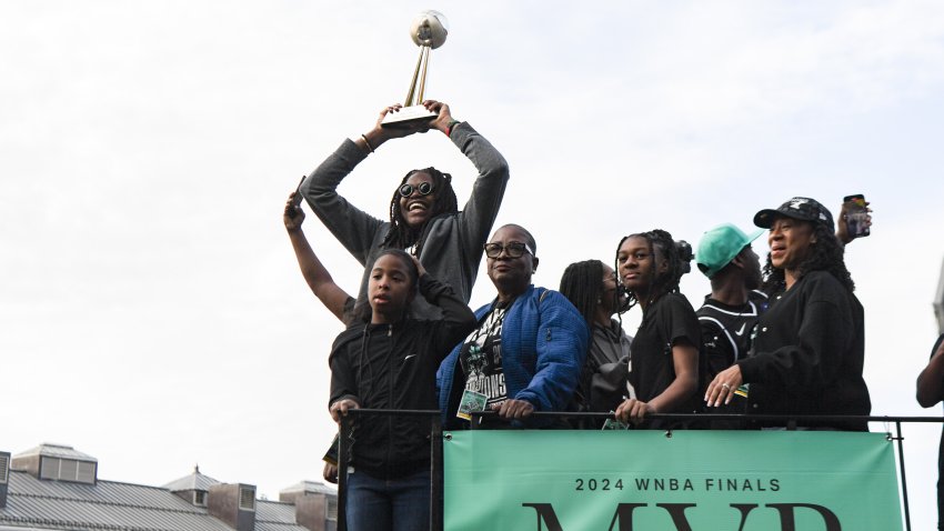 Jonquel Jones hoists the Finals MVP trophy above her head while riding on a float. She is surrounded by people. The float has a seafoam green banner on it that reads 2024 WNBA Finals MVP Jonquel Jones.